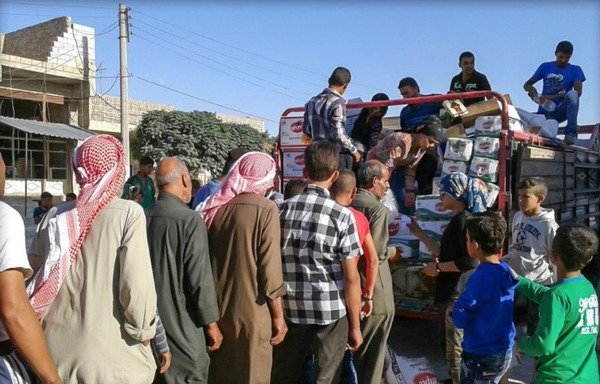 Des hommes représentants leurs ménages forment une ligne ordonnée pour recevoir l'aide humanitaire de la benne d'un véhicule de distribution du conseil militaire de Manbij. [Diyaruna]