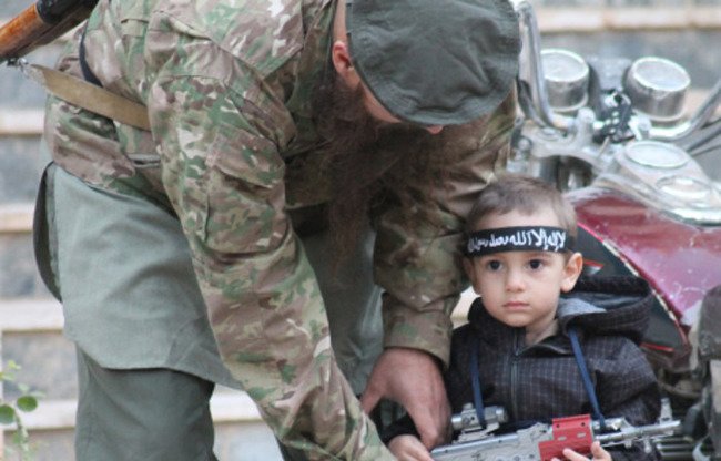 Wêneyeke cihê nîgeraniyê ku di babetekê de di dawîn kovara propagendayî ya DAIŞ, "Rûmiya", de hatiye belav kirin diyar dike çawa bavek kurê xwe fêrî hilgirtina çekekî dike. Zarok li deverên DAIŞ tên rahênankirin daku nifşê bê ji çekdaran pêk bînin.